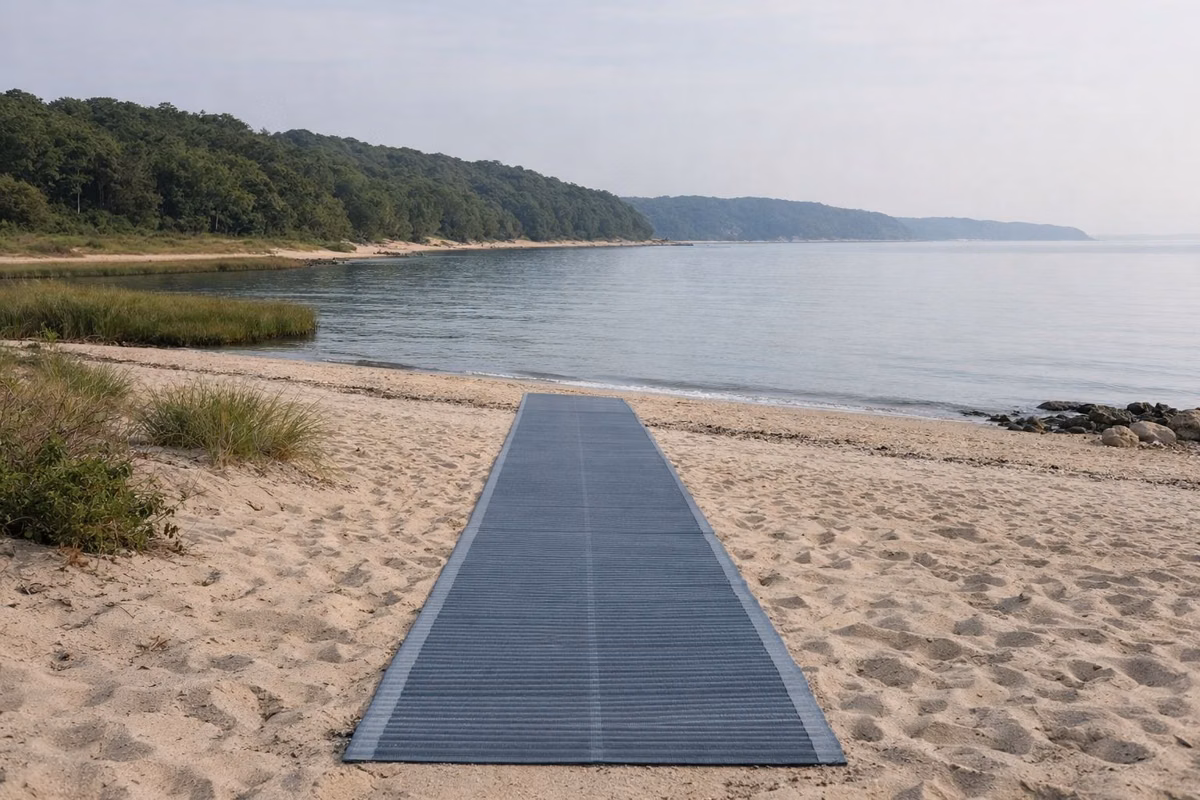 A blue-gray roll-out beach access mat laid across pale sand toward the water's edge, calm water beyond