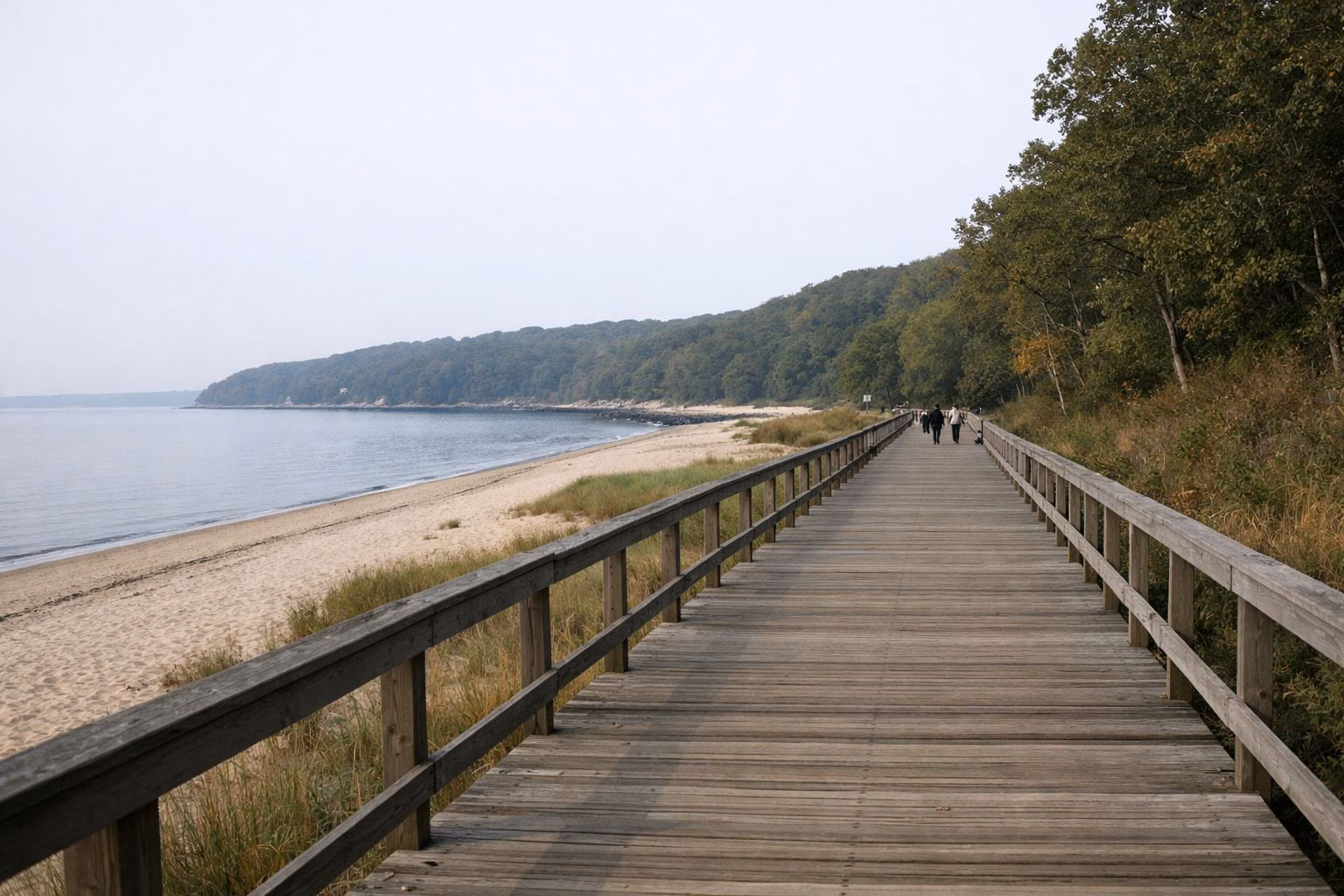 A long, wide wooden boardwalk running beside a sandy beach, a few walkers in the distance, calm sound on one side