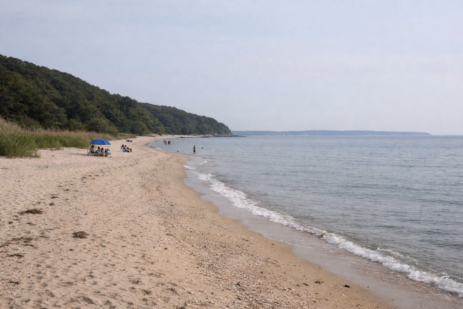 A wide, calm bay beach of pale sand on a sheltered sound, low gentle waves, a faint low coastline across the water on the horizon, a few distant beachgoers small in the frame
