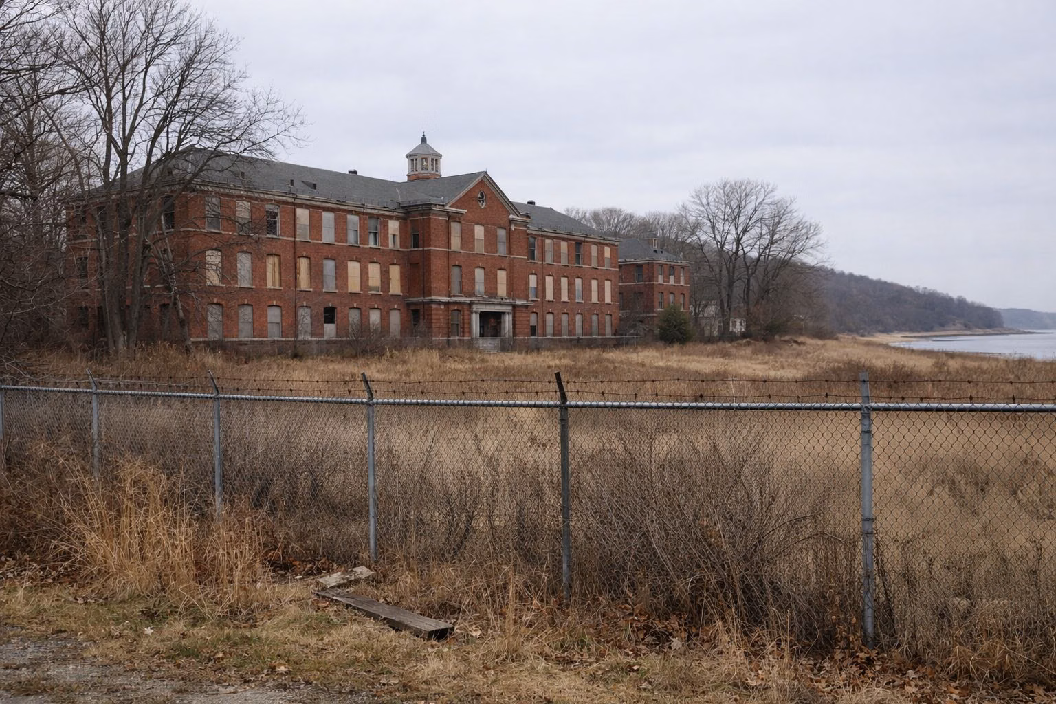 A large abandoned red-brick institutional building with boarded windows behind a chain-link fence, overgrown grass and bare trees