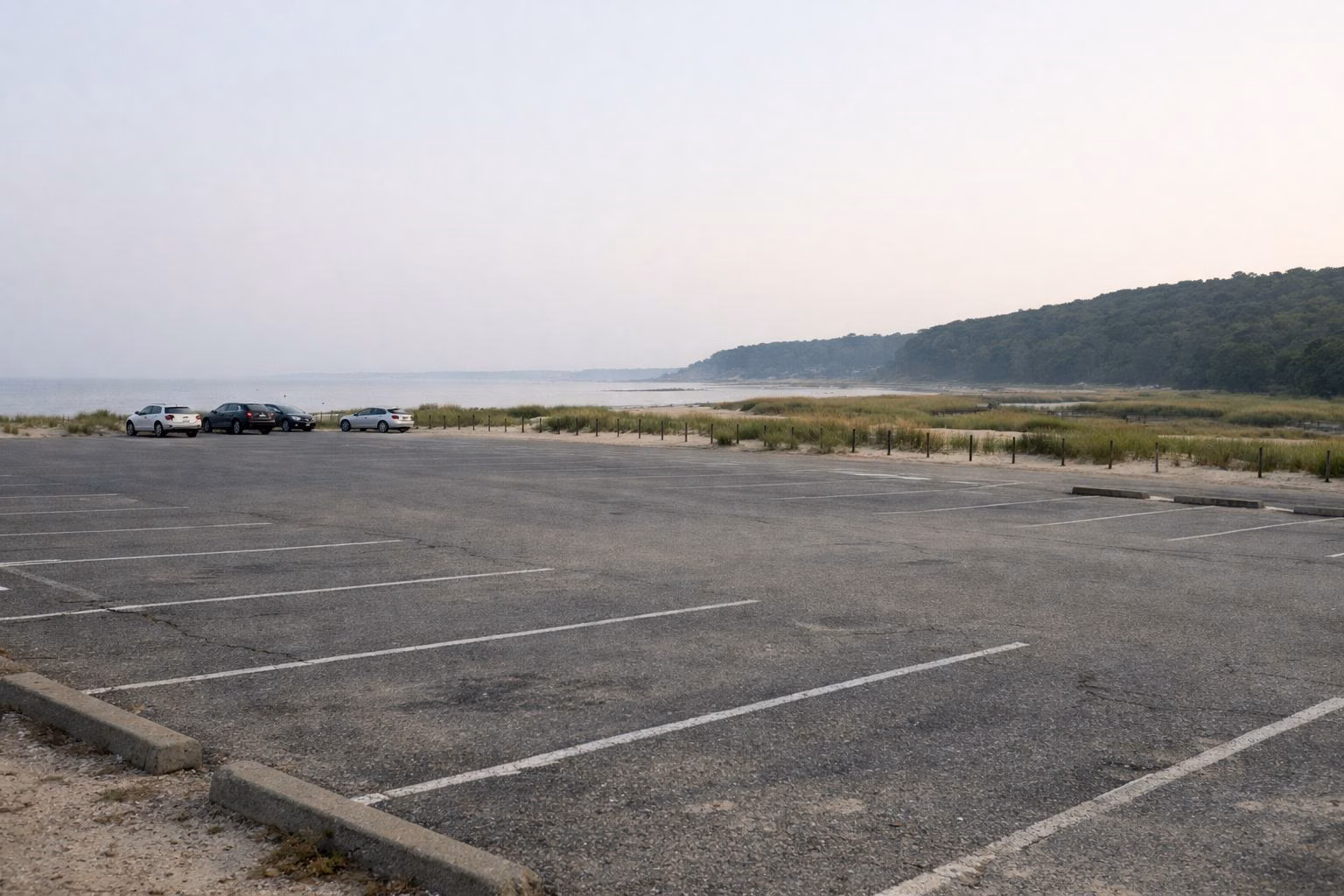 A large, mostly empty paved beach parking lot in early morning, a few parked cars far away, low dunes and dune grass beyond