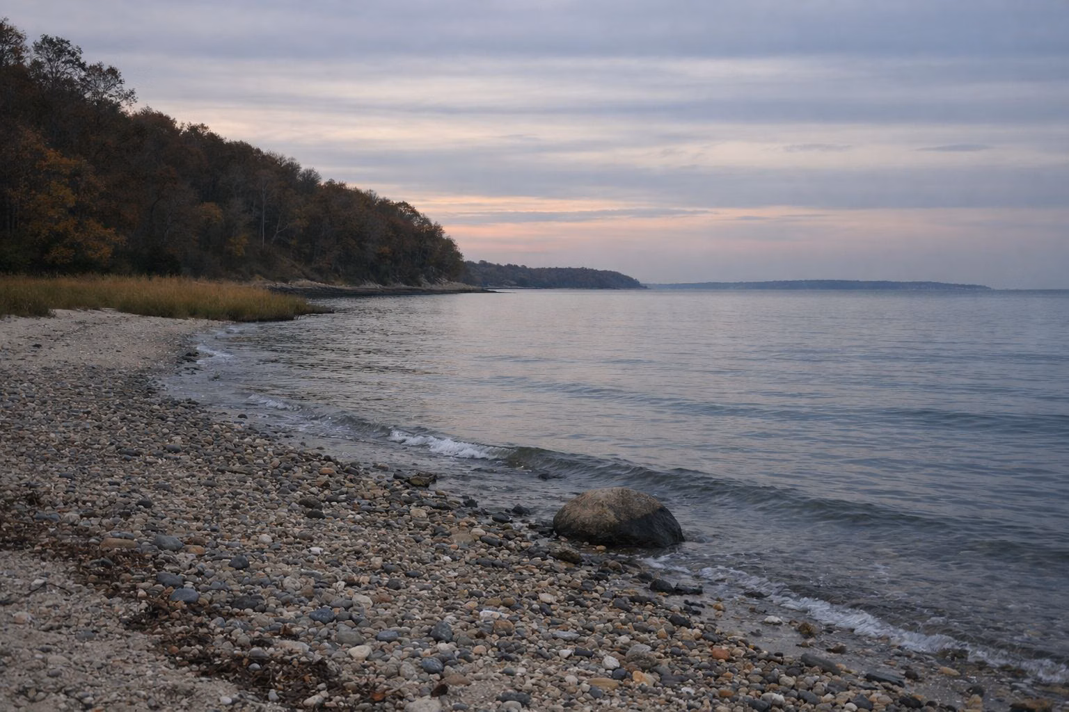 A pebbly shoreline on a calm sound at dusk, a faint coastline far across the water